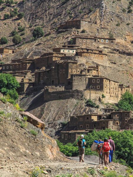 Im Hintergrund sieht man ein Berberdorf im Hohen Atlas, das terrassenförmig an den Berghang gebaut wurde. Im Vordergrund gehen mehrere Wanderreisende gerade auf das Dorf zu. Sie werden von einem Packesel begleitet, der ihre Ausrüstung trägt.