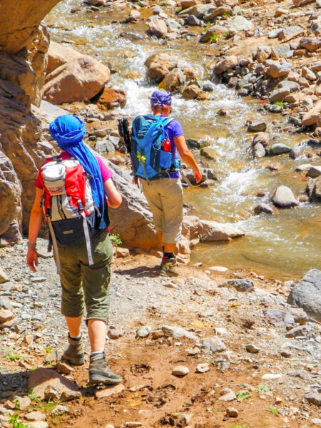 Zwei Personen auf Wanderreise folgen einem steinigen Pfad durch die Toubkalregion in Marokko auf einen schmalen Bach zu, den sie auf ihren Trekkingabenteuer queren müssen.