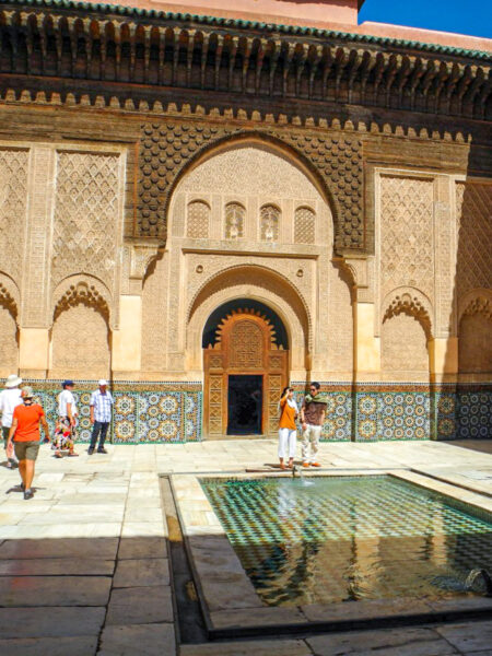 Mehrere Tourist*innen auf Wanderreise in Marokko besichtigen am Bild die Ben Youssef Madrasa, ein bekannte Koranschule in Marrakesch. Sie stehen im Innenhof des Gebäudes, in dessen Mitte sich ein Wasserbecken befindet.