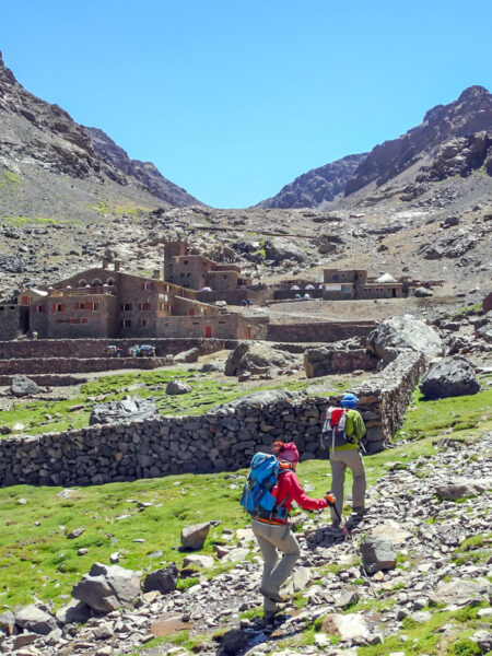 Zwei Wanderreisende gehen einen Pfad in Marokko entlang. Links von ihnen sieht man ein Steingebäude in der Region Toubkal.