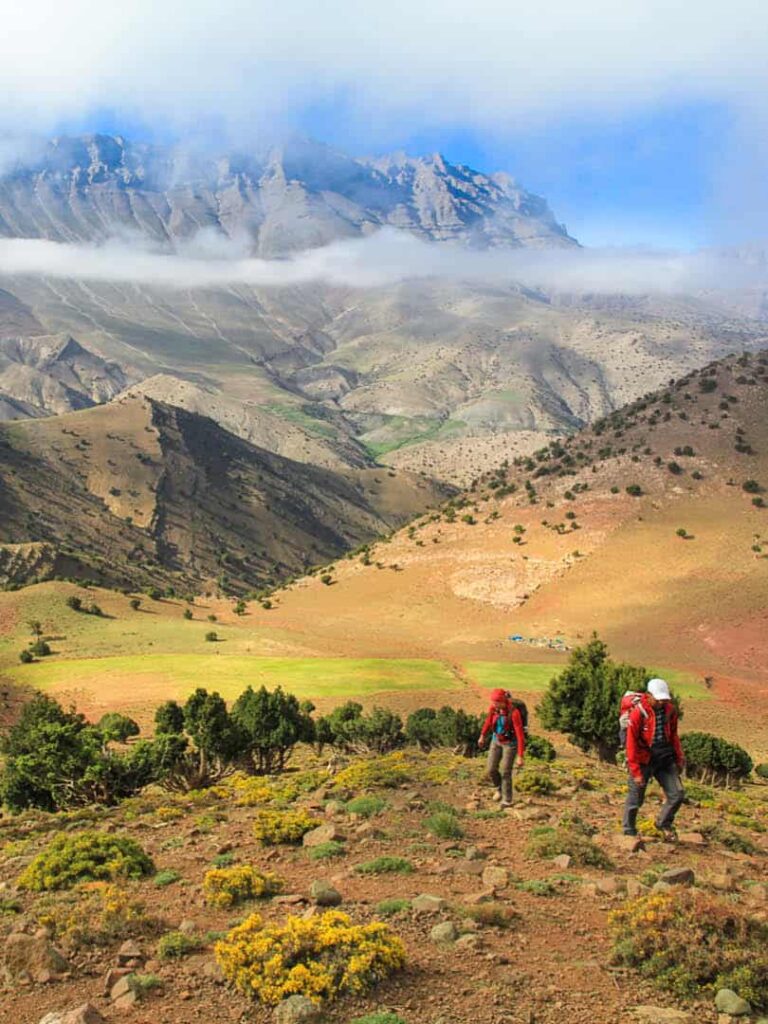Zwei Personen auf einer Wanderreise durch Marokko erklimmen einen Hang im Hohen Atlas. Hinter ihnen kann man das malerische Panorama der nordafrikanischen Gebirgskette ausmachen.