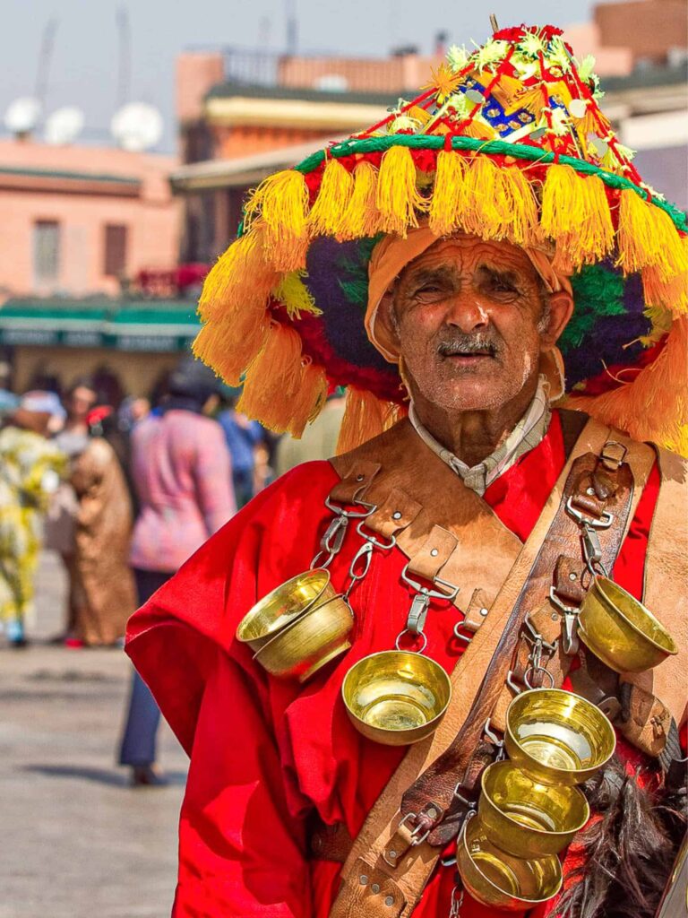 Auf dem Bild sieht man einen traditionellen Wasserträger in Marrakesch. Der ältere Mann trägt die traditionelle rote Uniform des Berufs, der heute vom Aussterben bedroht ist, historisch aber immens wichtig für die Infrastruktur in Marokko war.