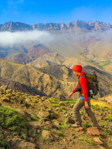 Eine lachende Frau auf einer Wanderreise geht durch unwegsames Gelände im Hohen Atlas. Hinter ihr breitet sich die Berglandschaft Marokkos aus, wo einzelne Wolkenschwaden zum Teil die Berge verdecken.