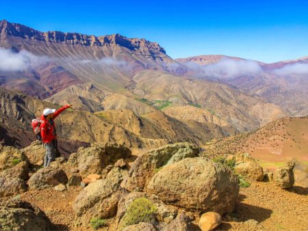 Ein Guide von Weltweitwandern deutet auf etwas in der Ferne im Hohen Atlas in Marokko. Hinter ihm sieht man die steilen Felswände der Region, die zum Teil hinter Wolkenschwaden verborgen sind.