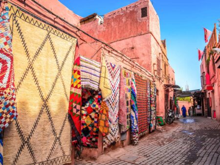 Wir sehen eine Straße in Marrakesch. Mehrere bunten Tücher oder Teppiche hängen vor einer der Wände herab und es scheint die Sonne.