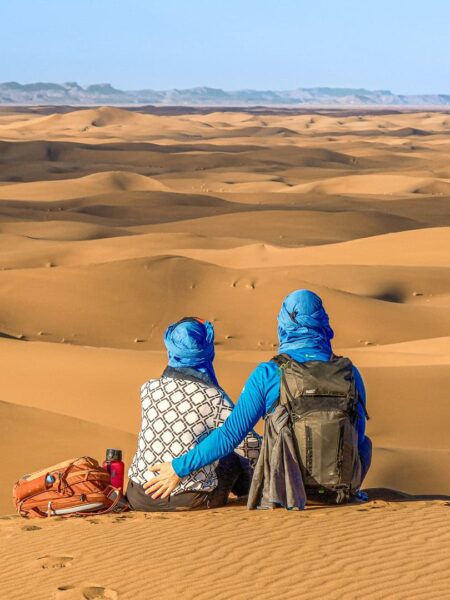 Ein Pärchen auf der Wanderreise in Marokko sitzt auf einer Sanddüne und schaut in die Wüste hinaus. Sie tragen blaue Kopftücher, um sich gegen die starke Sonne zu shcützen.