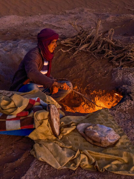 Ein Weltweitwandern-Guide kniet vor einem Feuer, das in einer Grube in der Wüste in Marokko brennt. Er versorgt die kleine Reisegruppe mit Essen.