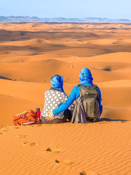 Zwei Personen auf Wanderreise sitzen Arm in Arm in der Wüste Marokkos. Sie beide tragen blaue Kleidung und Kopftücher.