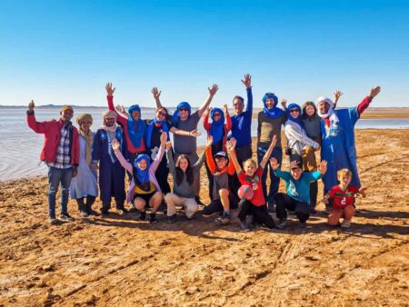 Auf dem Bild sieht man ein Gruppenfoto einer Familienreise und Wanderreise nach Marokko. Die Eltern und auch die Kinder jubeln in dem Bild auf dem Wüstensand.