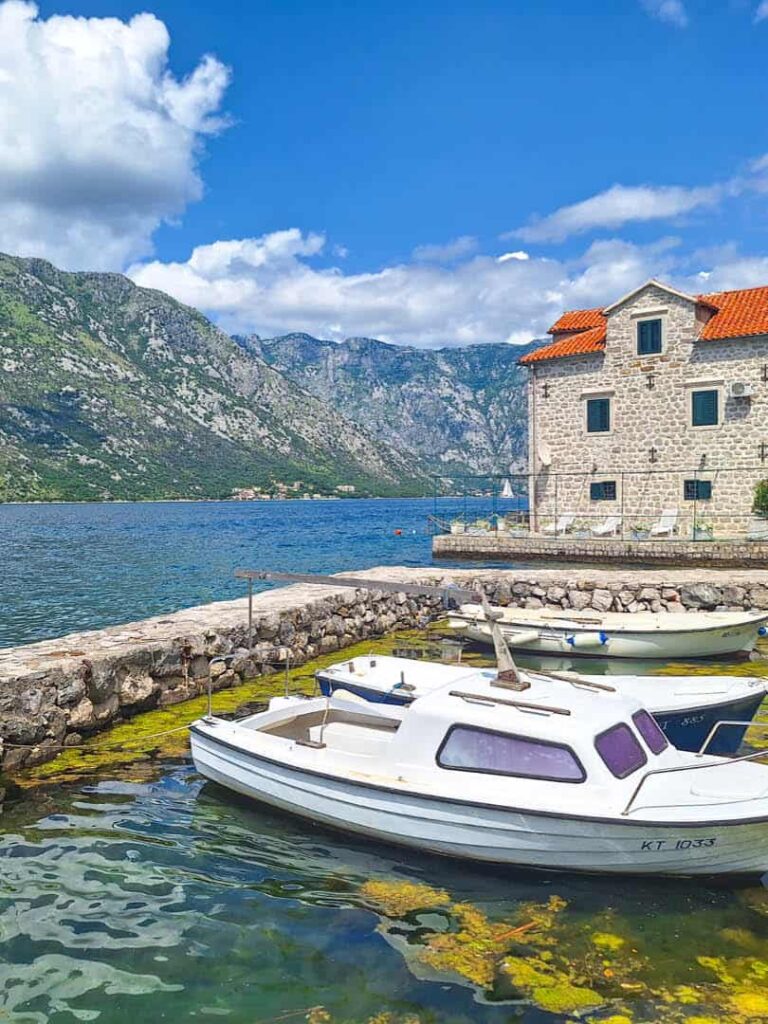 Wie sehen einen Ausschnitt der Bucht von Kotor. Im Vordergrund liegen zwei Boote in einem Hafen, während sich die Bucht in Montenegro im Hintergrund verliert.