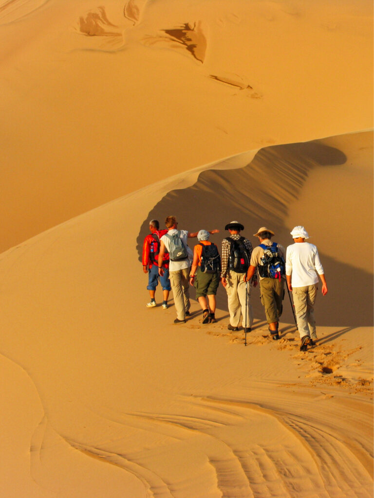Mehrere Personen auf einer Trekkingreise gehen hintereinander auf dem Grat einer Sanddüne in der Wüste Gobi.
