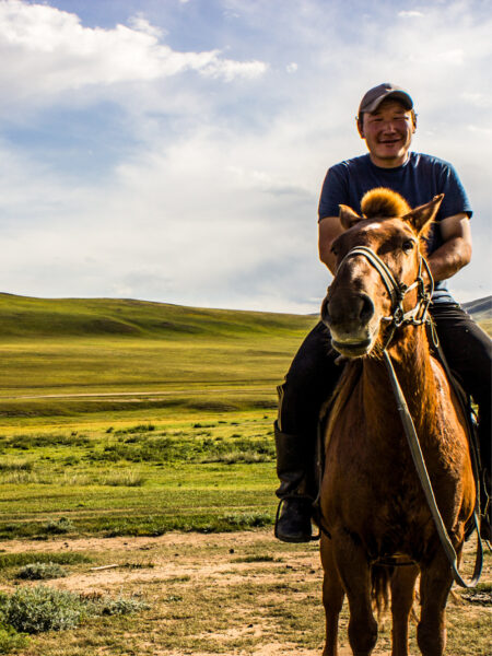 Ein Guide von Weltweitwandern sitzt auf einem Pferd und lacht in die Kamera. Hinter ihm kann man die Steppenlandschaft in der Mongolei sehen.