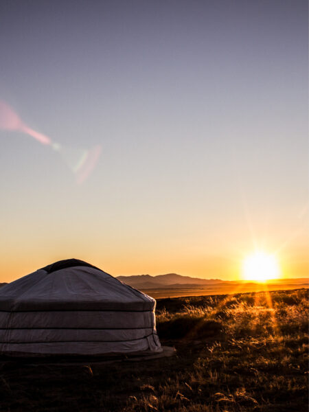 Die Sonne geht über einem Nomadencamp in der Mongolei unter. Im Vordergrund kann man eine der Jurten sehen, in der Reisegäste mit Weltweitwandern auf ihrem Trekkingurlaub durch die Mongolei übernachten.