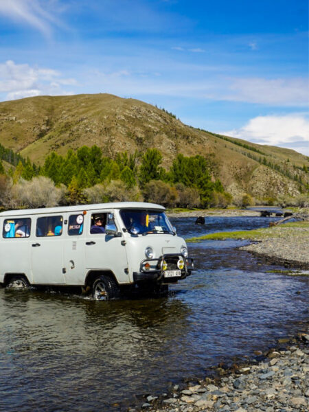 Ein weißer Transport mit der Reisesausrüstung einer Weltweitwandern-Trekking-Gruppe quert am Bild einen seichten Fluss.