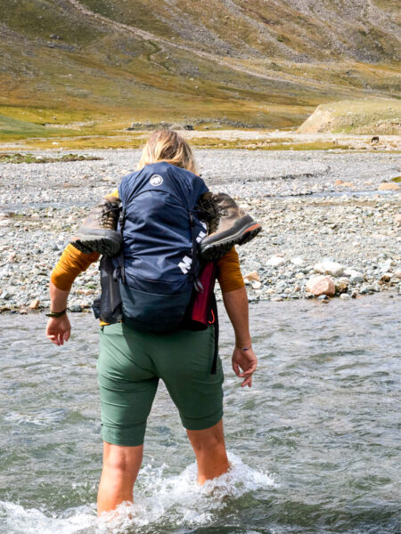 Eine Wanderin in der Mongolei passiert am Bild barfuß einen seichten Fluss. Die Schuhe hängen an ihrem Wanderrucksack, während sie durch die erfrischenden Fluten watet.