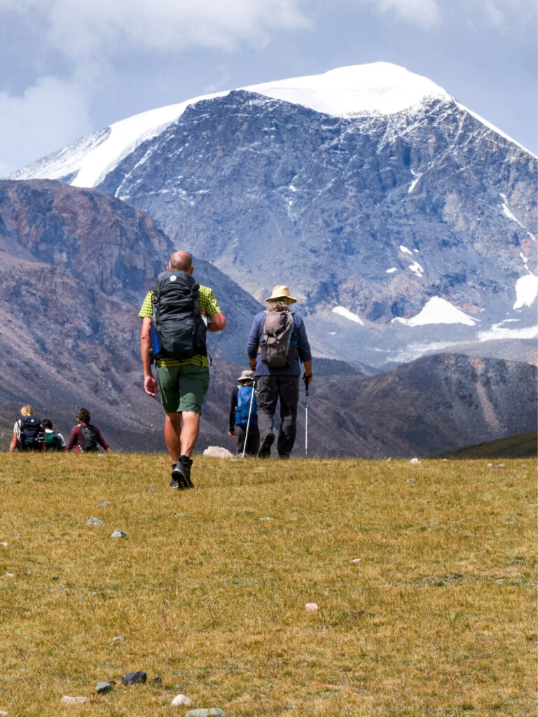 Zwei Personen auf einer Wanderreise durch den Westen der Mongolei gehen auf einen Berg im Hintergrund des Bildes zu. Bei dem Berg handelt es sich um einen Teil der Altai Sayan Gebirgskette.