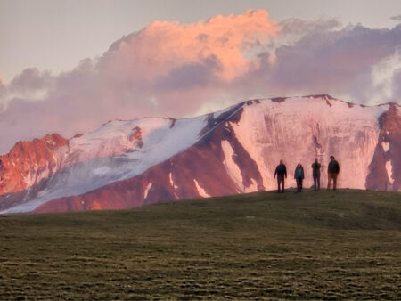 Mehrere Personen stehen auf einem Hügelkamm im Licht der untergehenden Sonne. Hinter ihnen sieht man den Gebirgszug der Altai Sajan in der Mongolei.