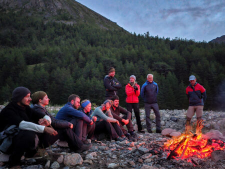 Eine Reisegruppe bei einer Trekkingreise durch die Mongolei sitzt bzw. steht rund um ein Lagerfeuer.