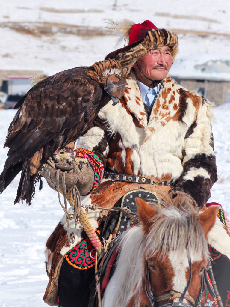 Ein kasachischer Adlerjäger in der Mongolei sitzt auf seinem Pferd vor einem schneebedeckten Hintergrund. Auf seiner Hand sitzt sein Adler. Das Foto wurde im Zuge des Adlerfestivals.