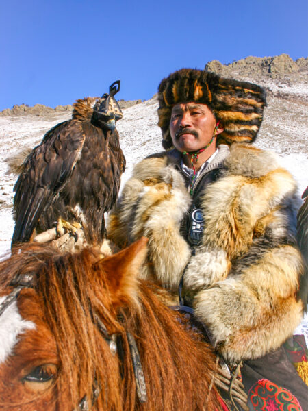 Ein mongolischer Reiter in traditioneller Kleidung mit diversen Fellen sitzt auf seinem Pferd vor einem schneebedeckten Hintergrund. Auf seiner Hand sitzt der Adler für das kommende Adlerfest, in dem er teilnimmt.
