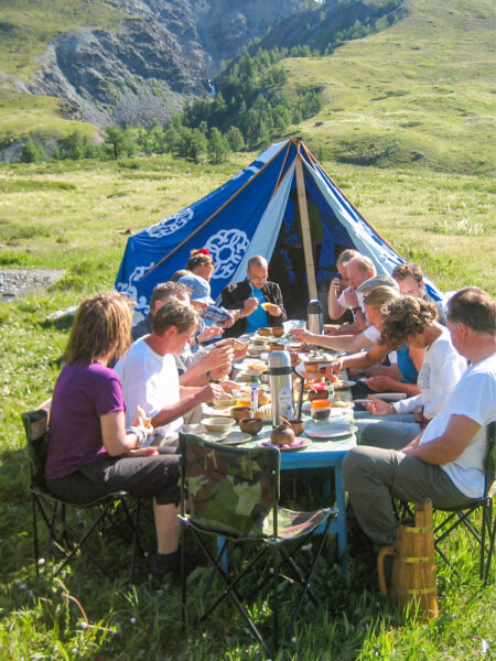 Eine Gruppe auf Wanderreise macht eine gemeinsame Essenspause vor ihrem Zelt. Hinter ihnen und um sie herum kann man Teile der mongolischen Gebirgslandschaft im Westen des Landes, nahe der Grenze zu China und Russland, sehen.