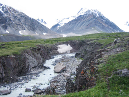 Ein Landschaftsfoto aus dem Altai Tavan Bogd Nationalpark in der Mongolei. Im Vordergrund sieht man einen kühlen Gebirgsbach, während im Hintergrund die schneebedeckten Gipfel der Region zu sehen sind.