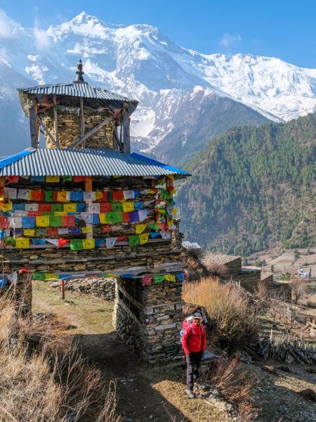 Ein Mann steht vor einem Tor auf dem Annapurna-Circuit-Trek in Nepal. Hinter ihm und dem großen Torgebäude kann man das Annapurna-Massiv sehen.
