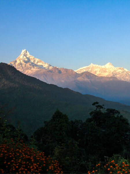 Eine Aussicht in Nepal. Im Fokus des Bildes liegt das Annapurna-Massiv im Himalaya-Gebirge in Nepal im Hintergrund. Im Vordergrund sind mehrere im Schatten liegenden Hügel zu erkennen.