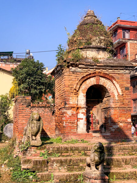 Auf dem Bild ist ein Tempel in Bhaktapur zu sehen. Er ist aus roten Ziegelsteinen gefertigt und mehrere kleine Statuen sind vor den Stufen zu sehen.