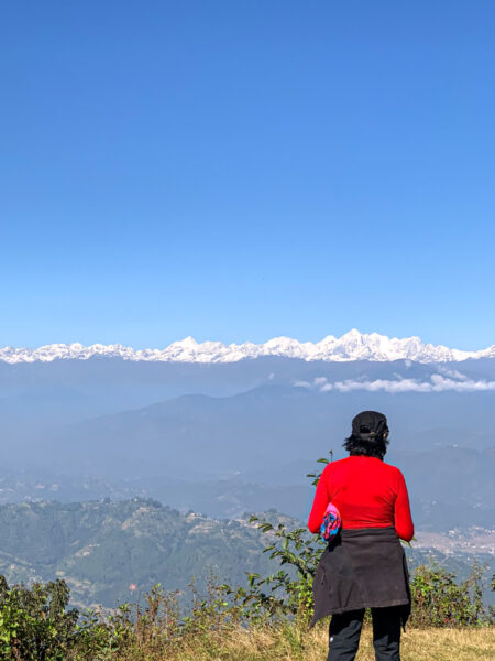 Eine Frau in einem roten Oberteil steht vor einer Aussicht in Nepal und schaut in die Richtung des Himalaya. Das wurde auf ihrer Wanderung nach Namobuddha aufgenommen.
