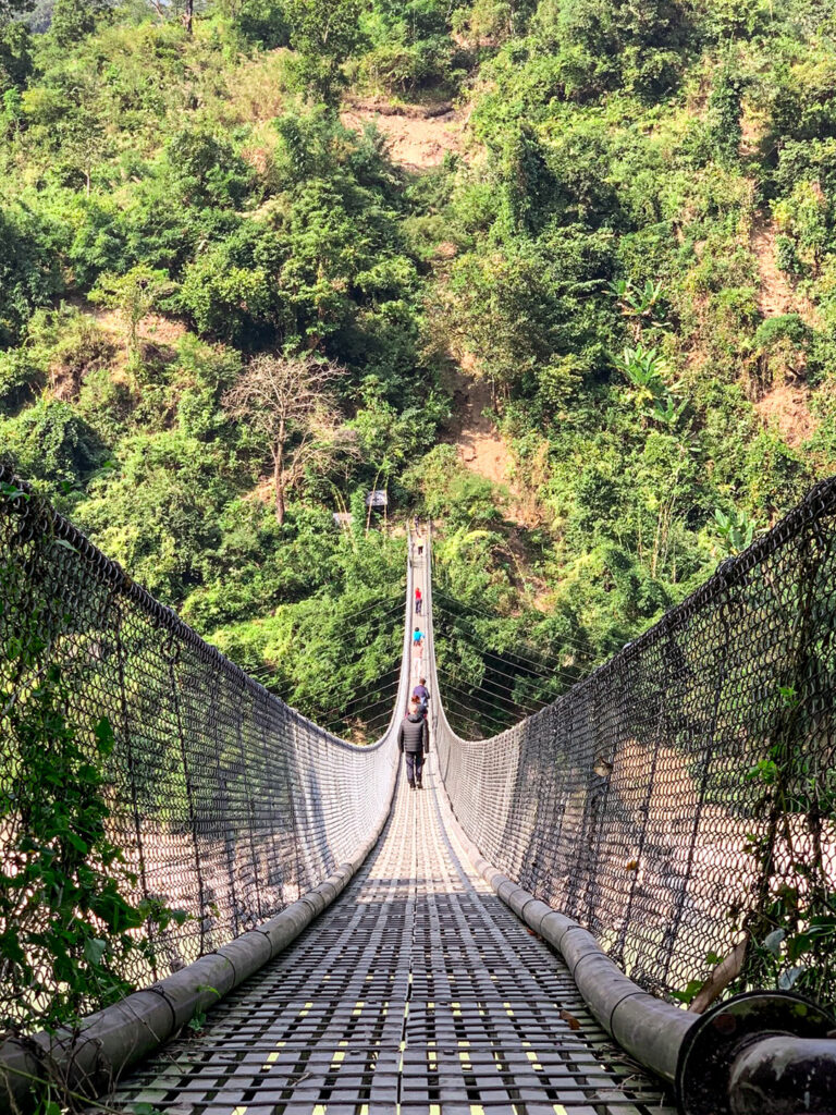 Man sieht eine Hängebrück in Nepal, die auf einen dichten Wald zuführt. Auf der Brücke stehen mehrere Personen auf einer Wanderreise durch das asiatische Land.