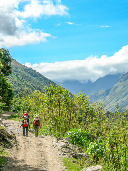 Drei Personen wandern am Salkantay-Trail in Peru, der sie nach Machu Pichu führt. Rechts neben dem Wanderweg führt ein Abhang hinnunter und im Hintergrund erkennt man das Bergland der Anden.