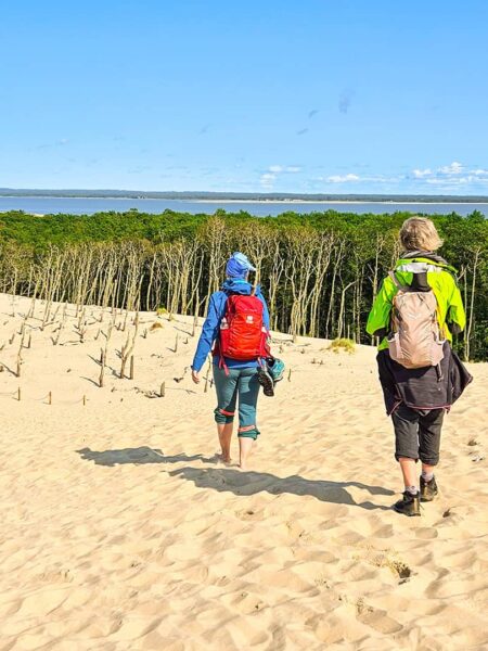 Zwei Personen gehen über die Dünen auf die Küste der Ostsee zu. Eine davon trägt einen roten Rucksack von Weltweitwandern und hält ihre Schuhe in der Hand, um barfuß durch den Sand zu spazieren, die andere Person geht ihr hinterher.