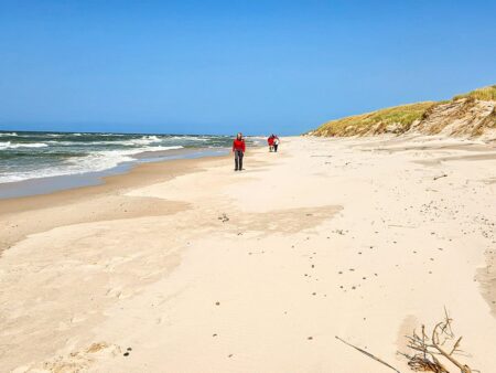 Zwei Personen in roten Jacken von Weltweitwandern stehen auf der Küste der Ostsee. Der Himmel über ihnen ist strahlend blau und die Wellen brechen links von ihnen auf dem Sand.