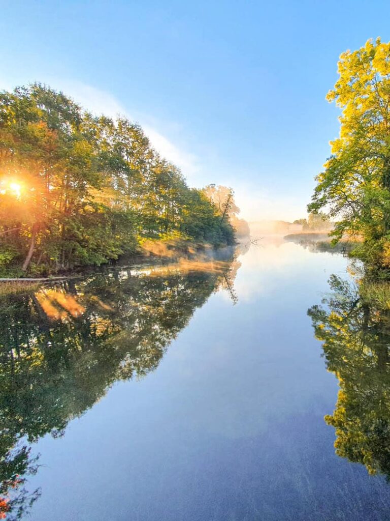Man sieht den Fluss Krutynia in Polen, im Licht der Morgensonne. Das Waser wirkt still und die Bäume spiegeln sich darin, bevor Wanderreisende von Weltweitwandern die Ruhe in ihren Paddelbooten unterbrechen werden.