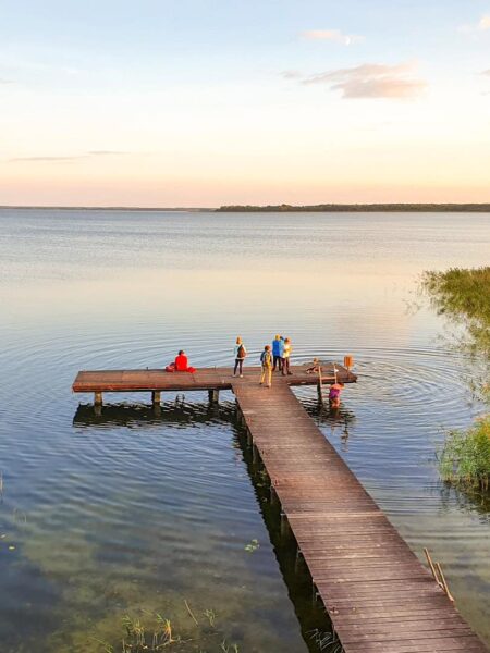 Drei Personen auf Wanderreise stehen auf einem Steg, der in einen polnischen See hineinragt. Eine weitere Person steigt gerade über eine Treppe ins Wasser hinab.