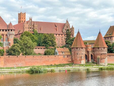 Man sieht die Marienburg in Malbork vom Wasser aus. Der rote Stein der Festung, die man auch im Zuge der geführten Reise mit Weltweitwandern ansehen kann, reflektiert leicht auf den Wellen.
