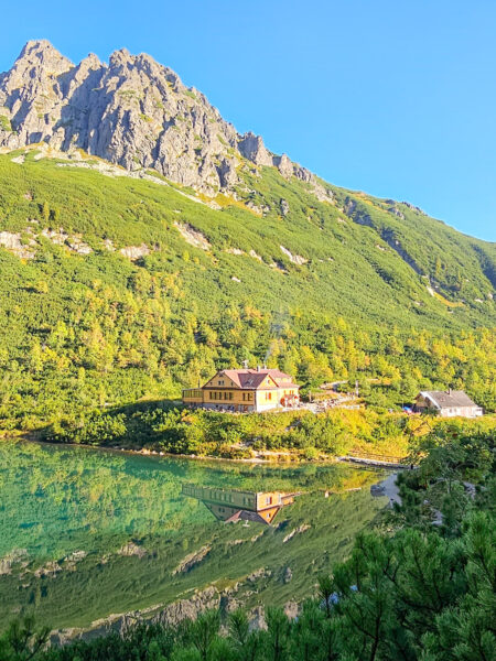Eine Berghütte am Grünen See oder Zelené Pleso in der Slowakei ist vom morgendlichen Lciht der Sonne angeschienen. Sie spiegelt sich im Wasser des bekannten Sees, von dem aus Wanderreisende mit Weltweitwandern in ihren Tag durch die Hohe Tatra in Polen starten.