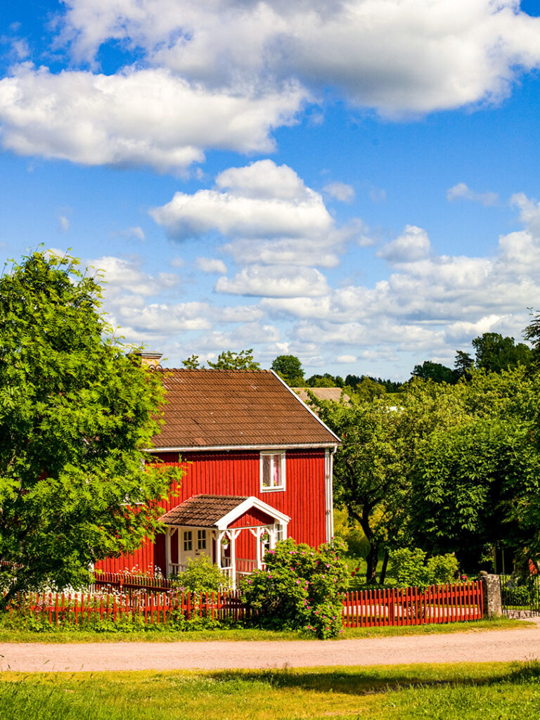 Ein rotes, schwedisches Haus steht neben einem unbefestigten Weg im Norden Schwedens. Ein paar Bäume verdecken es ein wenig.