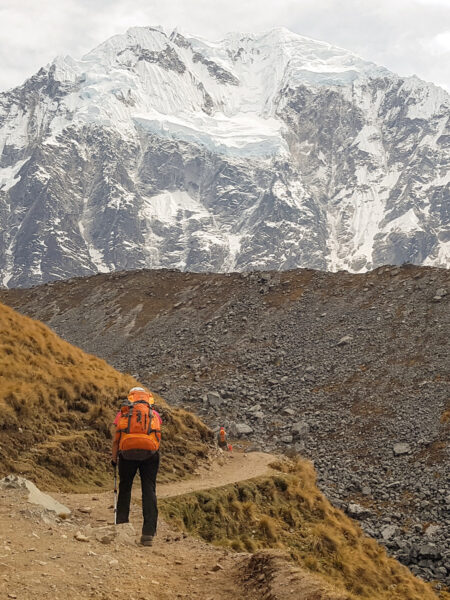 Eine Person mit einem großen Wanderrucksack marschiert auf ihrer Wanderreise über einen Wanderpfad in den Anden. Im Hintergrund sieht man einen schneebedeckten Gipfel.