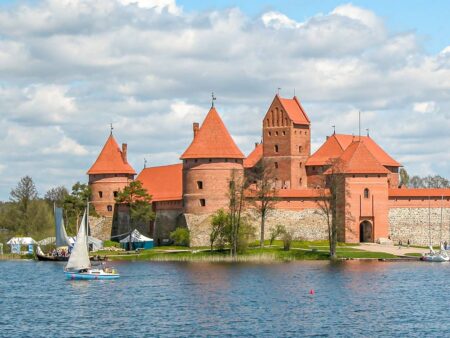 Die Wasserburg Trakai reflektiert am Wasser des Flusses über den hinweg man sie im Bild sehen kann. Ein kleines Segelboot hebt sich weiß vor den Zielgesteinen der Burg ab.