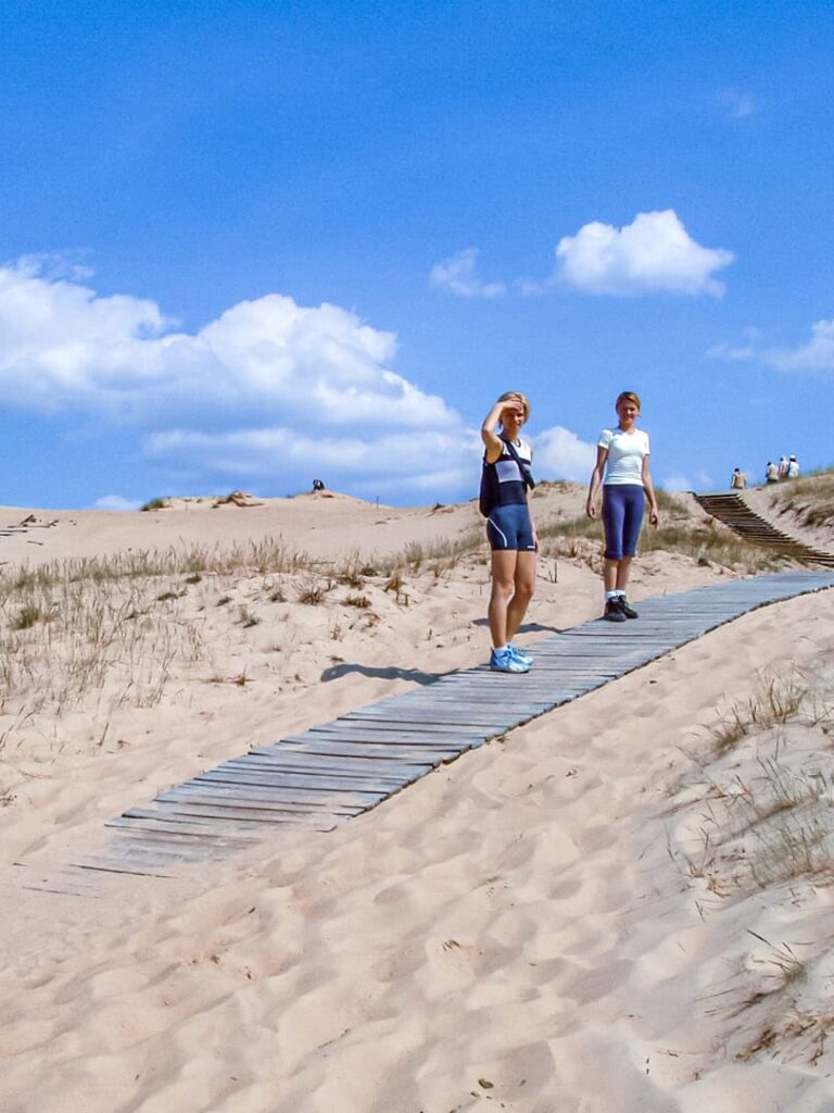 Zwei Frauen wandern über die toten Dünen in Litauen. Beide schaun in die Kamera und halten mit ihrer Hand die strahlende Sonne während ihrer Wanderung ab.
