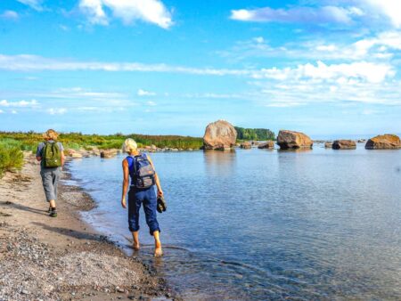 Zwei Personen auf Wanderreise gehen am Ufer des Pühajärv-Sees entlang. Beide tragen Rucksäcke und während am Ufer geht, hat sie die Schuhe ausgezogen und geht im Wasser des Sees herum.