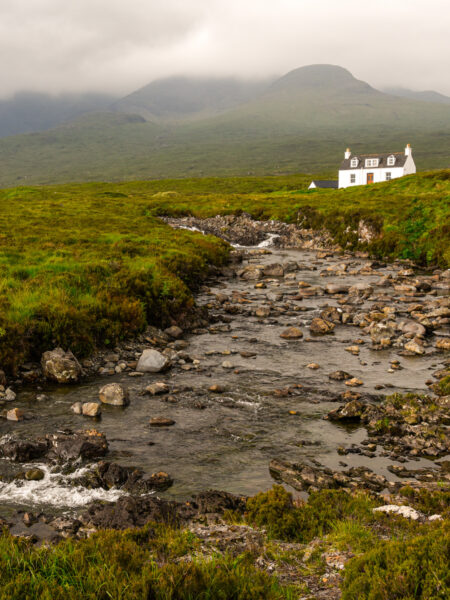 Ein Landschaftsfoto der Isle of Skye in Schottland. Hinter einem Bachlauf sieht man ein weißes Cottage, das Allt Dearg Cottage.