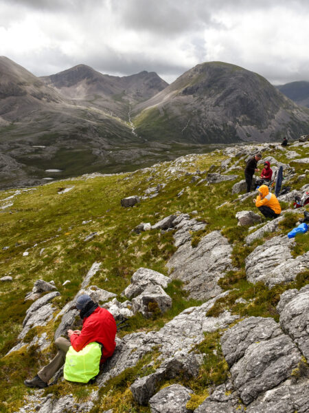 Mehrere Personen auf Wanderreise machen gerade eine Pause auf dem felsigen Untergrund in den schottischen Highlands. Im Hintergrund kann man einen Gebirgszug erkennen.