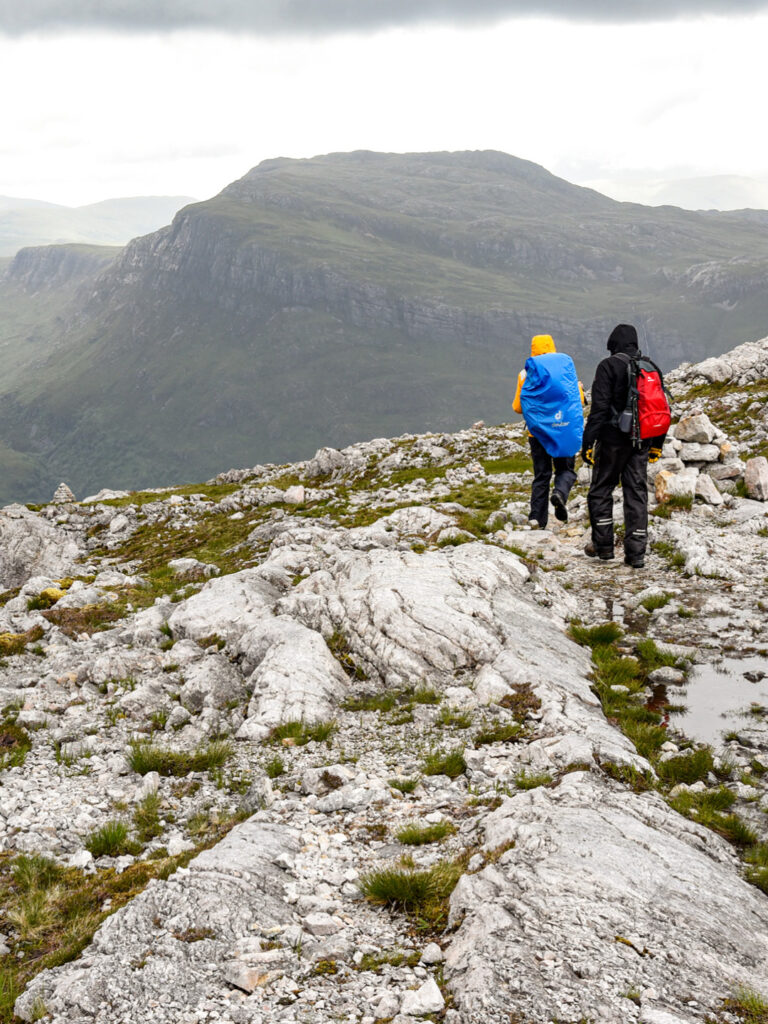 Zwei Wanderer folgen einem felsigen Pfad durch die schottischen Highlands. Sie tragen beide große Rucksäcke, die sie für die Wanderreise durch Schottland brauchen.