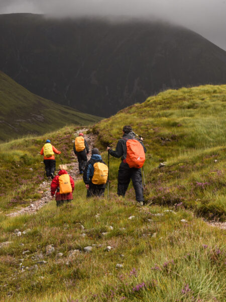 Ein kleine Gruppe aus fünf Personen auf Wanderreise marschiert durch die schottischen Highlands. Sie tragen Wanderrucksäcke und Ausrüstung gegen das feuchte Klima des Nordens.
