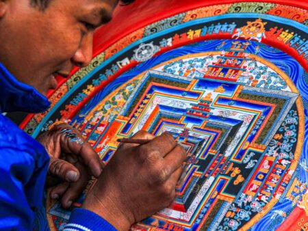 Ein nepalesischer Mandalakünstler beugt sich über sein Kunstwerk, ein Mandala, das Kreise und Quadrate sowie andere Muster verbindet und sowohl in rot als auch blau erstrahlt.