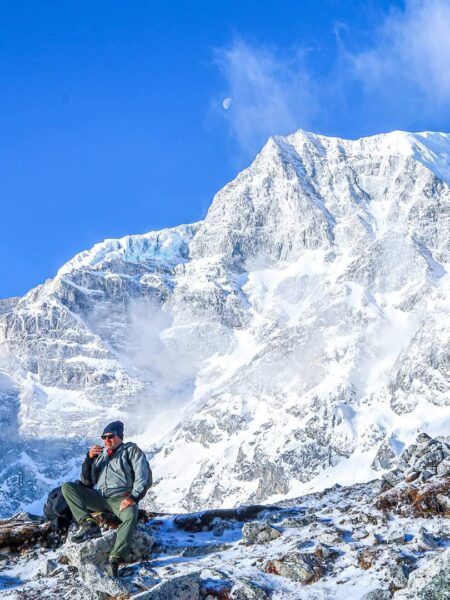 Im Vordergrund sitzt ein Wanderreisender mit Mütze und macht Pause. Im Hintergrund erhebt sich ein Berg in der Region Manaslu in Nepal.