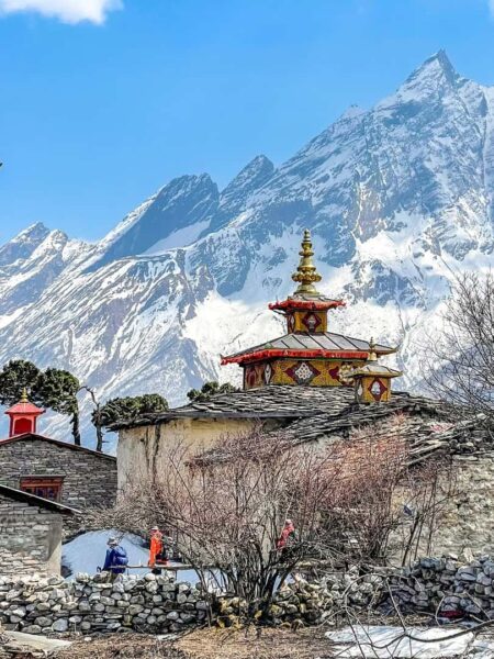 Eine kleine Gruppe auf Wanderreise geht durch in ein kleines Dorf in der Region Manaslu in Nepal. Man sieht im Hintergrund ein Bergmassiv, im Dorf erhebt sich das Dach eines kleinen Tempels oder Schreins neben den Reisenden.